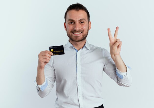 De glimlachende knappe mens houdt creditcard en gebaren twee met hand die op witte muur wordt geïsoleerd