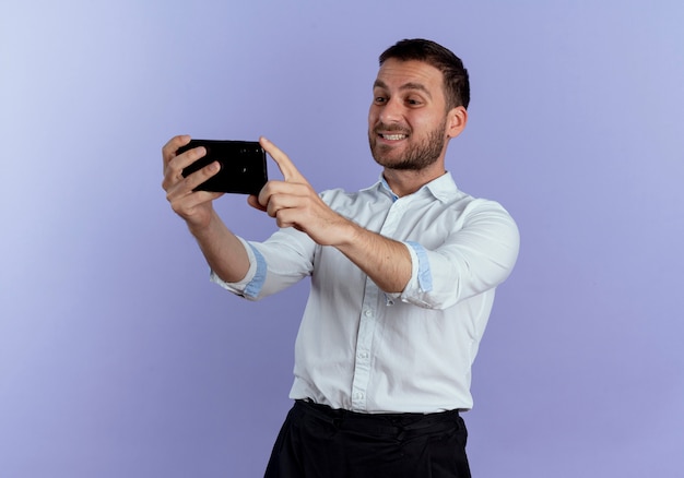 De glimlachende knappe mens houdt en bekijkt telefoon die op purpere muur wordt geïsoleerd