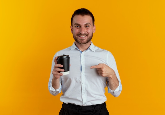 De glimlachende knappe mens houdt en wijst op koffiekop die op oranje muur wordt geïsoleerd