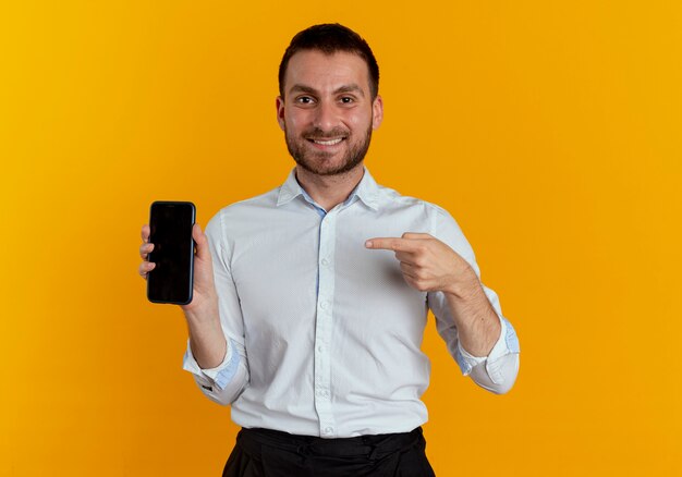 De glimlachende knappe mens houdt en wijst op telefoon die op oranje muur wordt geïsoleerd