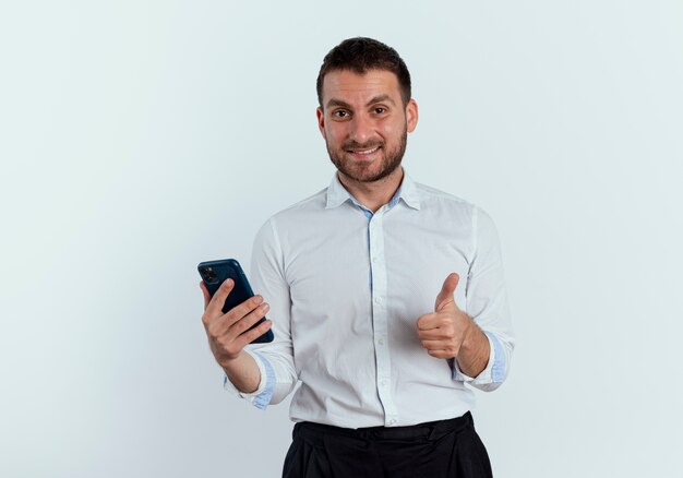 De glimlachende knappe mens houdt telefoon en duimen omhoog geïsoleerd op witte muur