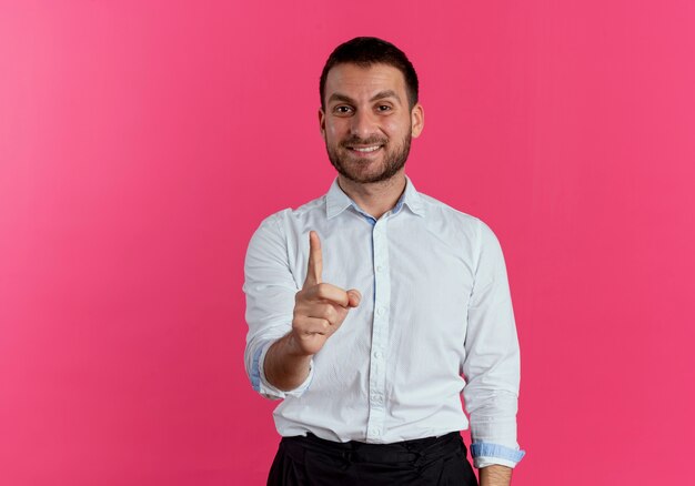 De glimlachende knappe mens toont wijsvinger die op roze muur wordt geïsoleerd