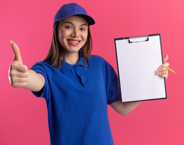 De glimlachende mooie bezorgvrouw in uniform houdt klembord en duimen omhoog geïsoleerd op roze muur met exemplaarruimte