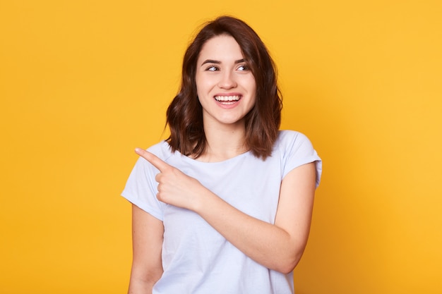 De glimlachende mooie jonge vrouw draagt opzij witte toevallige t-shirt, punten met wijsvinger