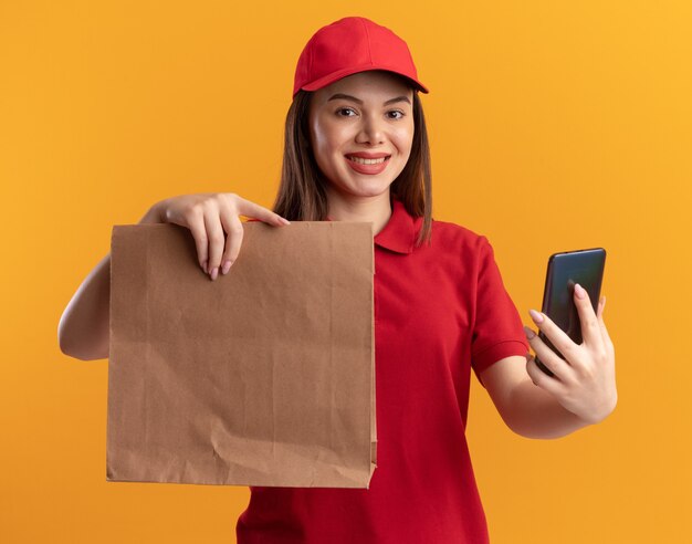 De glimlachende mooie leveringsvrouw in uniform houdt document pakket en telefoon op oranje