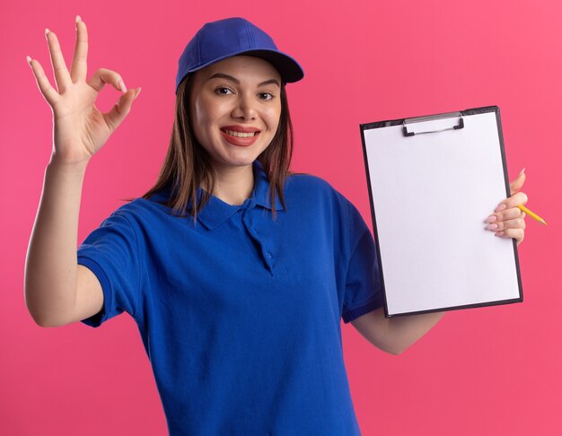 De glimlachende mooie leveringsvrouw in uniform houdt klembord en gebaren ok handteken dat op roze muur met exemplaarruimte wordt geïsoleerd