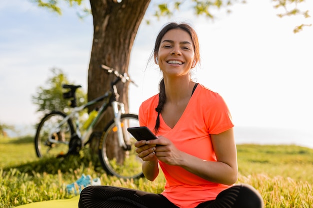 De glimlachende mooie telefoon van de vrouwenholding die sporten in ochtend in park doet