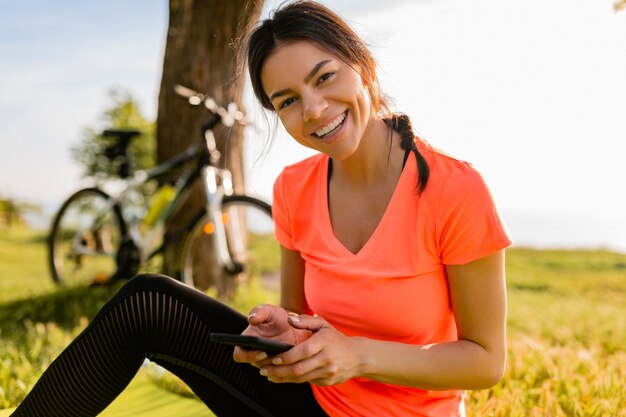 De glimlachende mooie telefoon van de vrouwenholding die sporten in ochtend in park doet