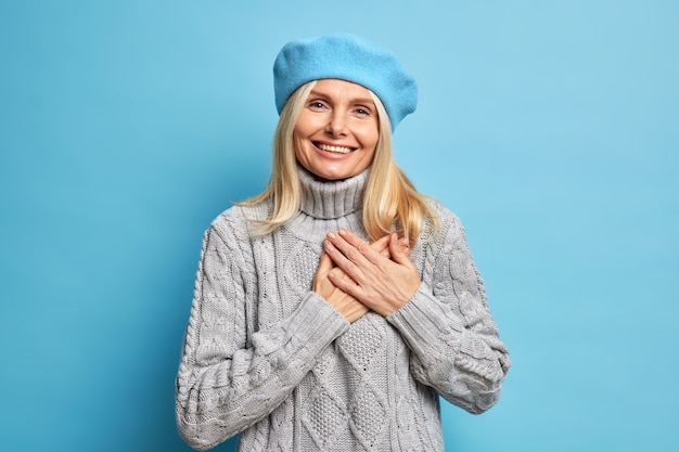 De glimlachende mooie vrouw houdt de handen tot het hart gedrukt drukt zijn dankbaarheid uit, draagt een blauwe baret en een gebreide grijze trui is dankbaar en waardeert uw hulp.