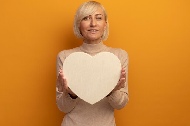 Gratis foto de glimlachende vrij blonde slavische vrouw houdt hartvorm die op oranje muur wordt geïsoleerd