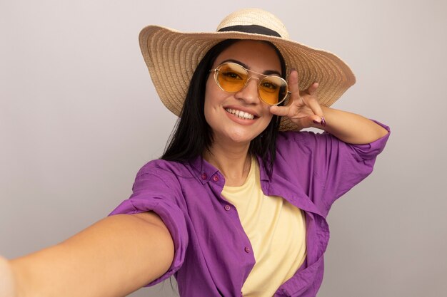 De glimlachende vrij donkerbruine vrouw in zonnebril met strandhoed gebaren drie met vingers pretendeert voor het nemen van selfie te houden die op witte muur wordt geïsoleerd