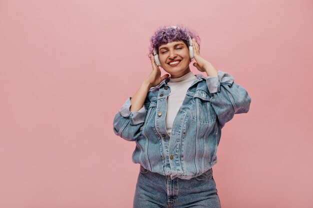 De glimlachende vrouw in blauw spijkerjasje luistert naar muziek met zijn ogen dicht. Vrouw in witte koptelefoon poseren op roze.