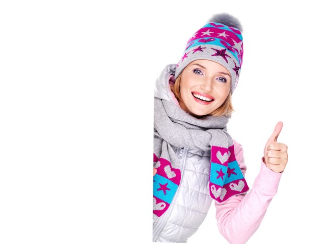 De glimlachende vrouw in de winterbovenkleding houdt het aanplakbiljet met duimen omhoog teken