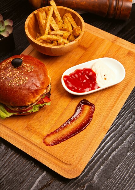 De hamburger van het kippenvlees met binnen tomaat en sla en frieten diende met zwarte olijf en ketchup op een houten dienblad