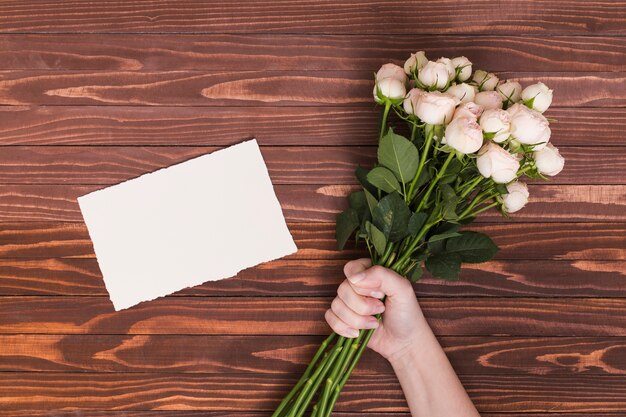 De hand die van de persoon bos van witte rozen houdt; blanco papier over houten bureau