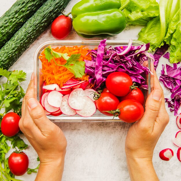De hand die van de persoon verse groenten en ingrediënten voor salade in container houdt