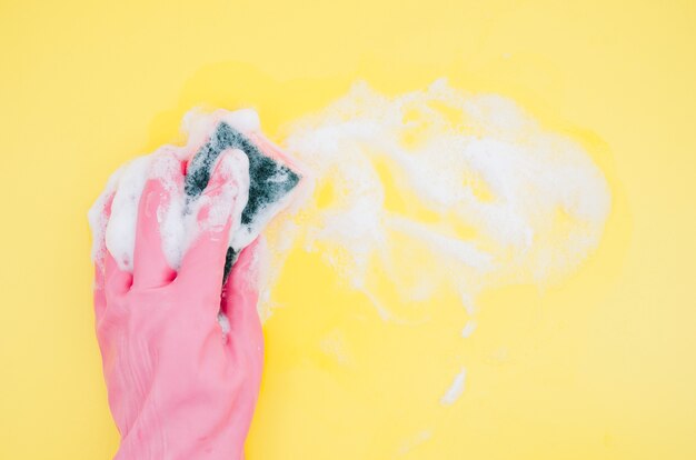 De hand die van een persoon roze handschoenen draagt die gele achtergrond met spons schoonmaken