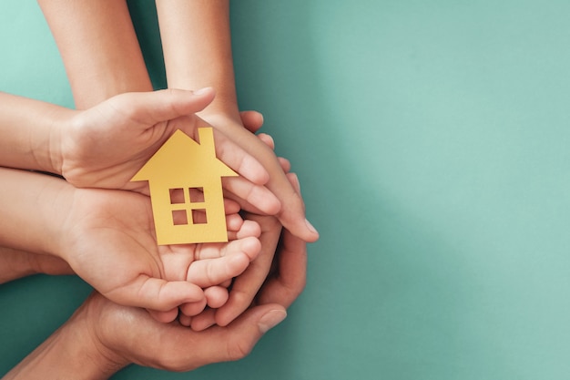 De handen die geel document huis op blauwe oppervlakte houden, familiehuis, dakloze schuilplaatshuisvesting en huis die verzekering, hypotheekconcept beschermen, bevorderen thuiszorg