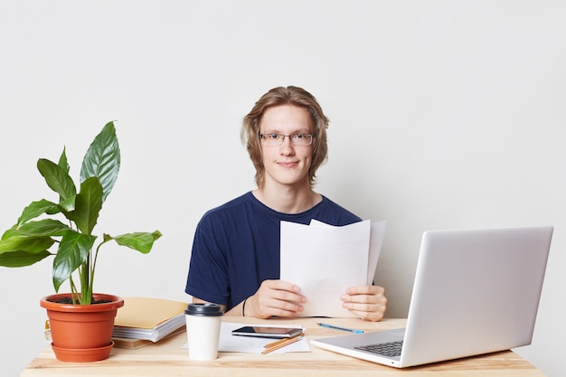 De hardwerkende professionele bedrijfsmedewerker zit op de werkplek, bekijkt zijn rekeningen, bestudeert documenten, met prachtige uitdrukking, gebruikt moderne technologieën voor werk