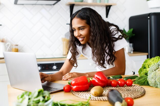 De jonge Afrikaanse vrouw typt iets in laptop op een keukenbureau met groenten