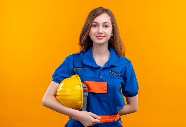 De jonge arbeider van de vrouwenbouwer in de veiligheidshelm die van de bouw eenvormige holding zelfverzekerd zich over oranje muur bevinden kijken