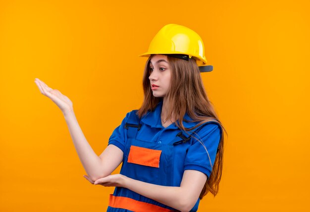 De jonge arbeider van de vrouwenbouwer in eenvormige bouw en veiligheidshelm die opzij kijken ontevreden opheffend hand die status over oranje muur opstelt