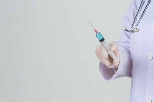 De jonge arts houdt hypodermische spuit met de rubberhandschoenen van het vaccinflesje op grijze muur.