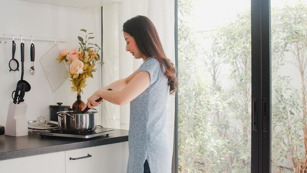 De jonge Aziatische Japanse dame geniet van thuis koken. Levensstijlvrouwen gelukkig het voorbereiden van voedsel die deegwaren en spaghetti voor ontbijtmaaltijd maken in moderne keuken bij huis in de ochtend.