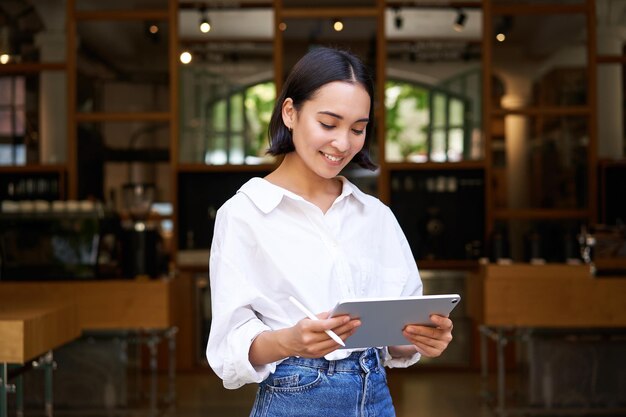De jonge Aziatische manager van de serveersterskoffie die met tablet en grafische tablet werkt