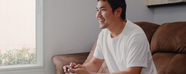 De jonge Aziatische mens die bedieningshendel het spelen videospelletjes in televisie in woonkamer gebruiken, mannetje die het gelukkige voelen voelen ontspant tijd thuis liggend op bank. Mannen spelen ontspannen thuis.