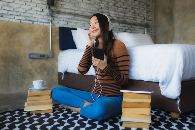 De jonge Aziatische vrouw die slimme mobiele telefoon met hoofdtelefoon met behulp van voor luistert muziek rond koffiekop en boek in slaapkamer