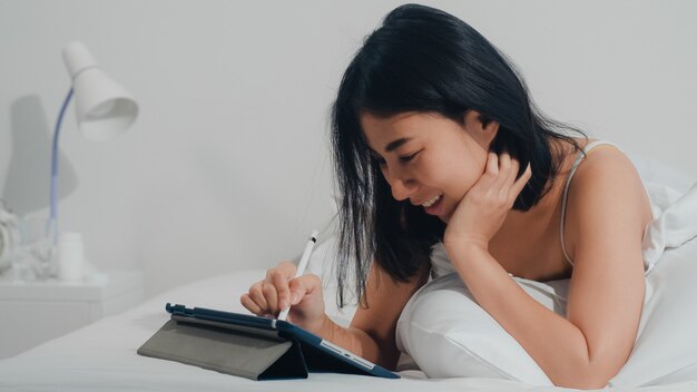 De jonge Aziatische vrouw die tablet gebruiken die sociale media controleren die het gelukkige glimlachen voelen terwijl het liggen op bed na kielzog omhoog thuis in de ochtend, het Aantrekkelijke Indische vrouwelijke glimlachen ontspant thuis in slaapkamer.