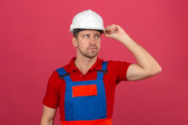 De jonge bouwersmens in eenvormige bouw en veiligheidshelm denken met verwarde hand op hoofd kijkt het hebben van twijfels over geïsoleerde roze muur