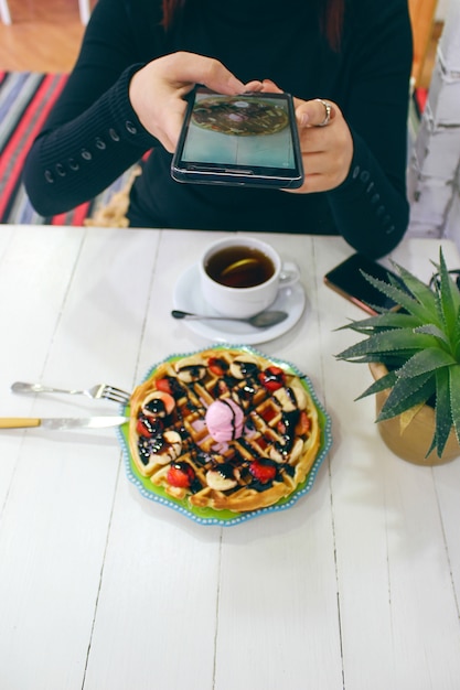 De jonge cafe die van de meisjeszitting ontbijtwafel met chocoladesaus, banaanplakken en aardbeien eten op groene ceramische plaat en fotografeerde haar ontbijt