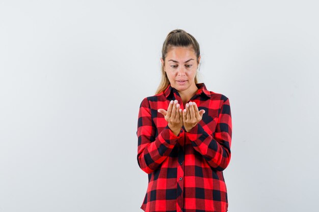 De jonge dame die haar tot een kom gevormde handen in casual overhemd bekijkt en verbaasd kijkt. vooraanzicht.