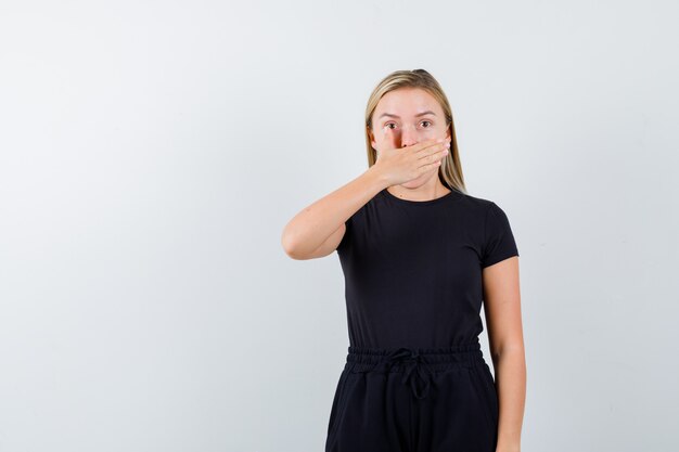 De jonge dame die mond behandelt met hand in t-shirt, broek en bang kijkt. vooraanzicht.