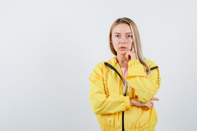 De jonge dame die zich in het denken bevindt stelt in t-shirt, jasje en kijkt zelfverzekerd