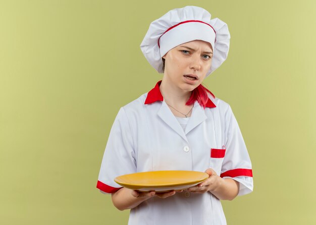 De jonge geërgerde blonde vrouwelijke chef-kok in eenvormige chef-kok houdt plaat en kijkt geïsoleerd op groene muur