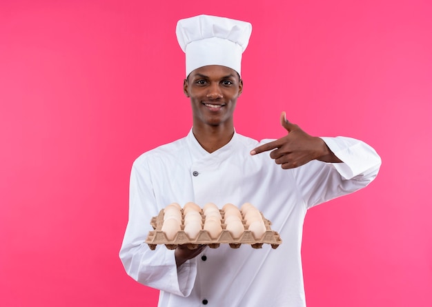 De jonge glimlachende Afro-Amerikaanse kok in eenvormige chef-kok houdt en wijst op partij verse eieren die op roze muur wordt geïsoleerd