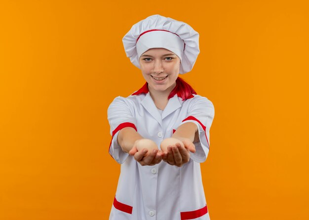 De jonge glimlachende blonde vrouwelijke chef-kok in eenvormige chef-kok houdt eieren uit geïsoleerd op oranje muur