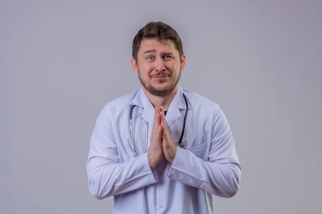 De jonge hoopvolle ongerust gemaakte arts die witte laag dragen en de stethoscoopholding dienen in bidden