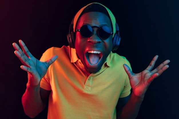 De jonge knappe blij verrast hipster man luisteren muziek met koptelefoon in zwarte studio met neonlichten.