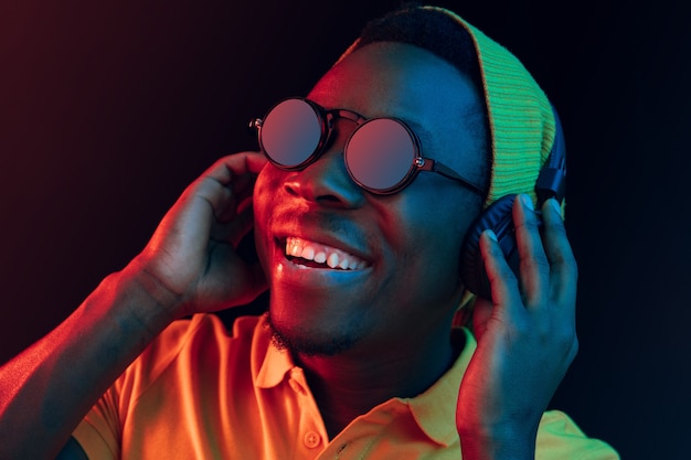 De jonge knappe gelukkig hipster man luisteren muziek met koptelefoon in zwarte studio met neonlichten. Disco, nachtclub, hiphopstijl, positieve emoties, gezichtsuitdrukking, dansconcept