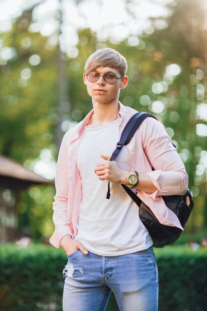 De jonge knappe man in vrijetijdskleding in zonnebrillen en een rugzak staat op de campus