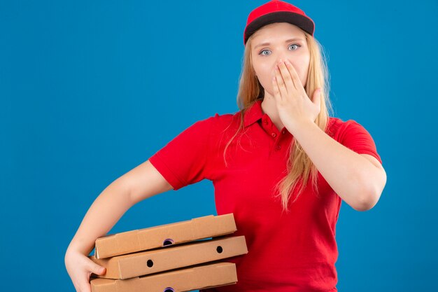 De jonge leveringsvrouw die rood poloshirt en GLB dragen die zich met geschokte pizzadozen bevinden die mond behandelen met overhandigen geïsoleerde blauwe achtergrond