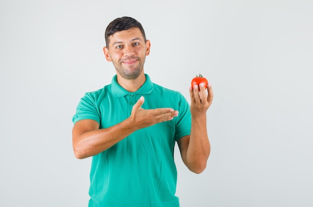 De jonge mens die tomaat in zijn hand in groenachtig t-shirt toont en gelukkig kijkt.