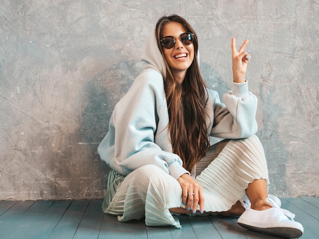 De jonge mooie glimlachende vrouw toont vredesteken. Trendy meisje in casual zomer hoodie en rok kleding.