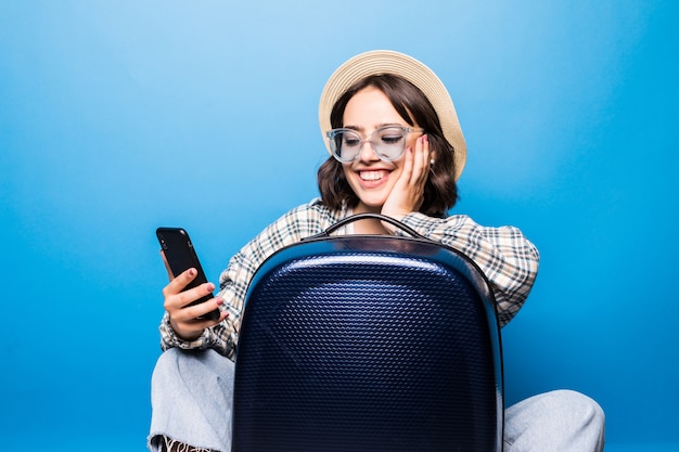 De jonge mooie vrouw in zonnebril en strohoed met een koffer onderzoekt de geïsoleerde telefoon. Vlucht reizen.