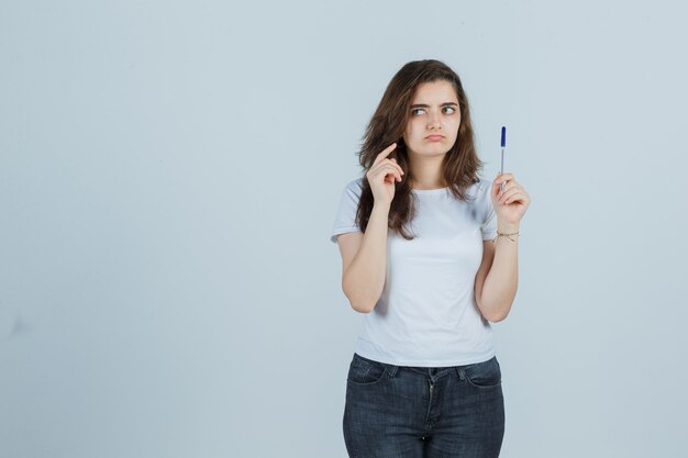 De jonge pen van de meisjesholding terwijl het denken in t-shirt, jeans en nadenkend kijkt. vooraanzicht.