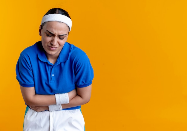De jonge pijnlijke Kaukasische sportieve vrouw die hoofdband en polsbandjes draagt houdt buik op sinaasappel met exemplaarruimte